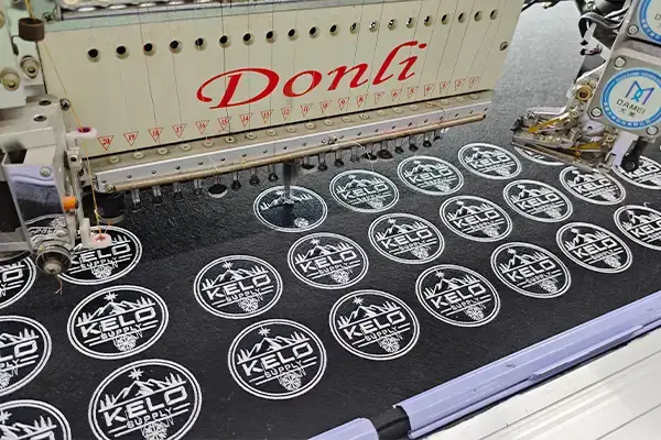 Multi-head embroidery machine producing repeated circular logo patches with consistent stitch detail on black fabric.