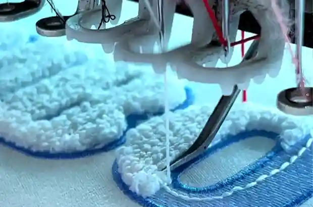 Close-up of an industrial embroidery machine stitching white chenille and blue embroidered details on a custom letter patch.