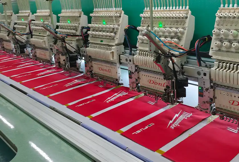 Multi-head industrial embroidery machines stitching white logos on red handbag fabric panels during bulk production.
