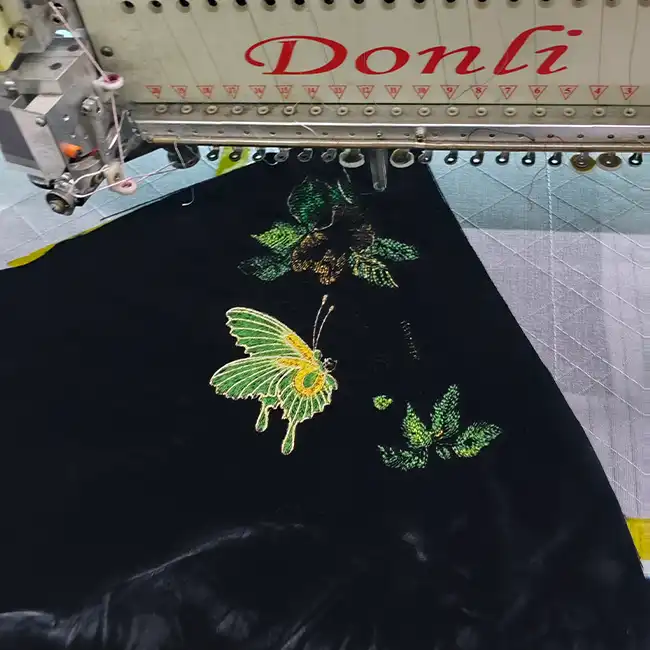 Industrial embroidery machine stitching a detailed butterfly and green floral design on a black fashion garment panel.