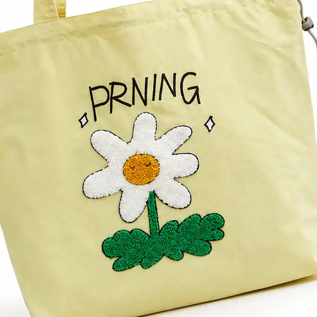 Light yellow handbag featuring chenille embroidery with a white daisy, green leaves, and black text on the front panel.