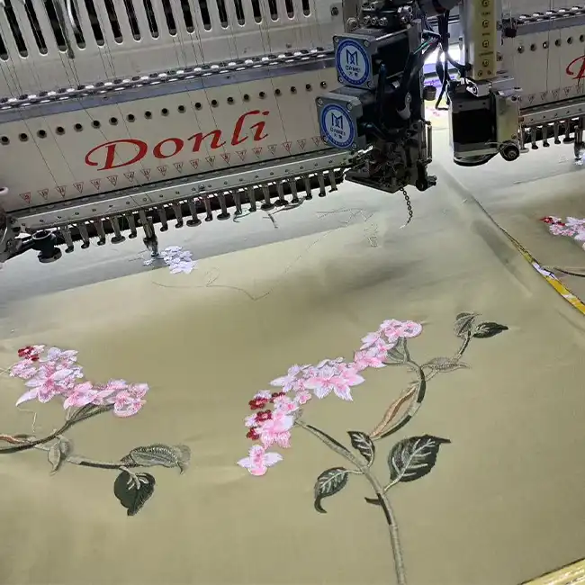 Industrial embroidery machine stitching delicate pink floral and green leaf designs on a light-colored fashion garment panel.