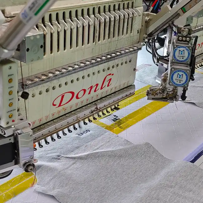 Industrial multi-head embroidery machine positioned over a gray garment panel during precision logo embroidery production.