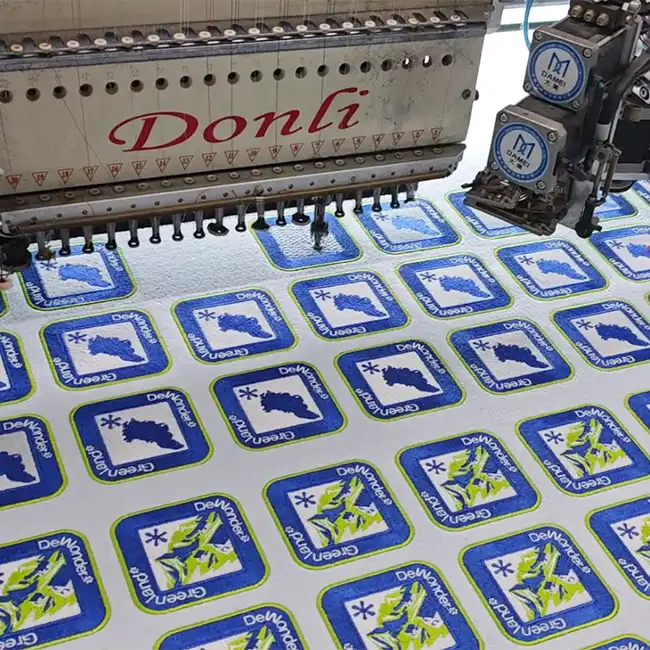 Industrial embroidery machine producing custom embroidered labels for handbags in bulk with blue and green border designs.