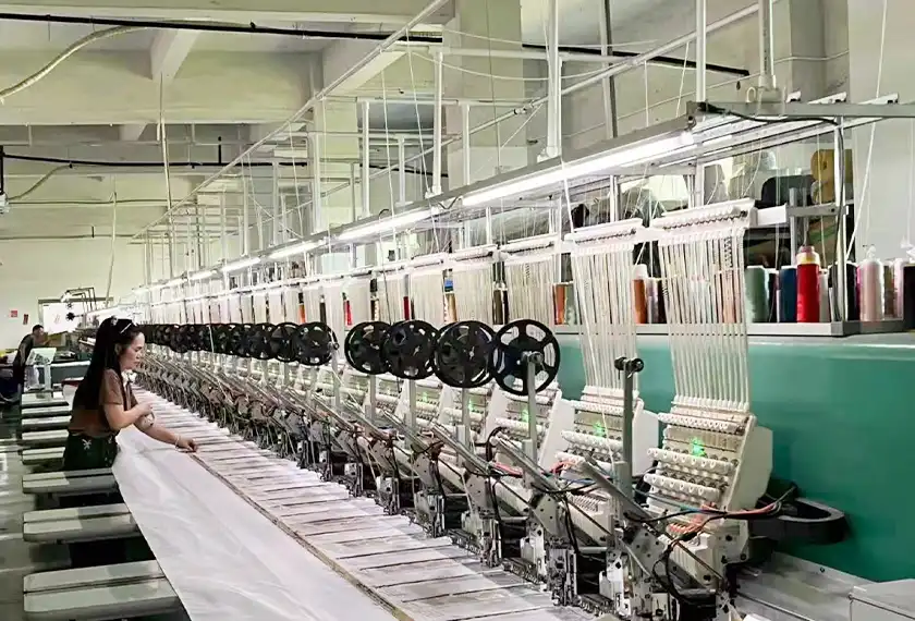 Long multi-head embroidery production line with a worker handling garment panels during bulk embroidery manufacturing.