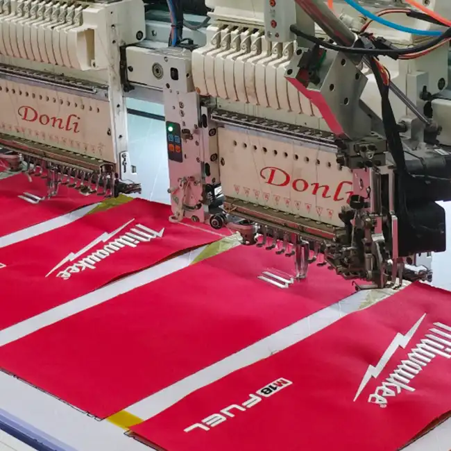 Industrial multi-head embroidery machines stitching white logo designs on red handbag fabric panels during bulk production.